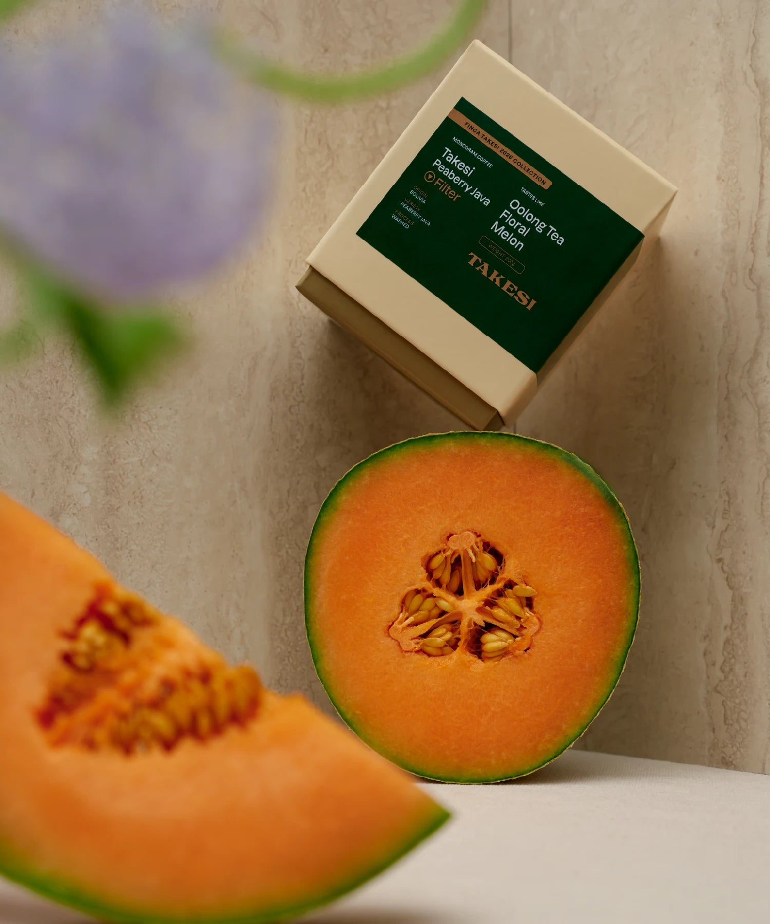 A cream and dark forest green box of Monogram Takesi Peaberry Java filter coffee resting on a surface in a still life setup, framed by a vibrant, cut-open cantaloupe melon showing its orange flesh and seeds in the foreground.