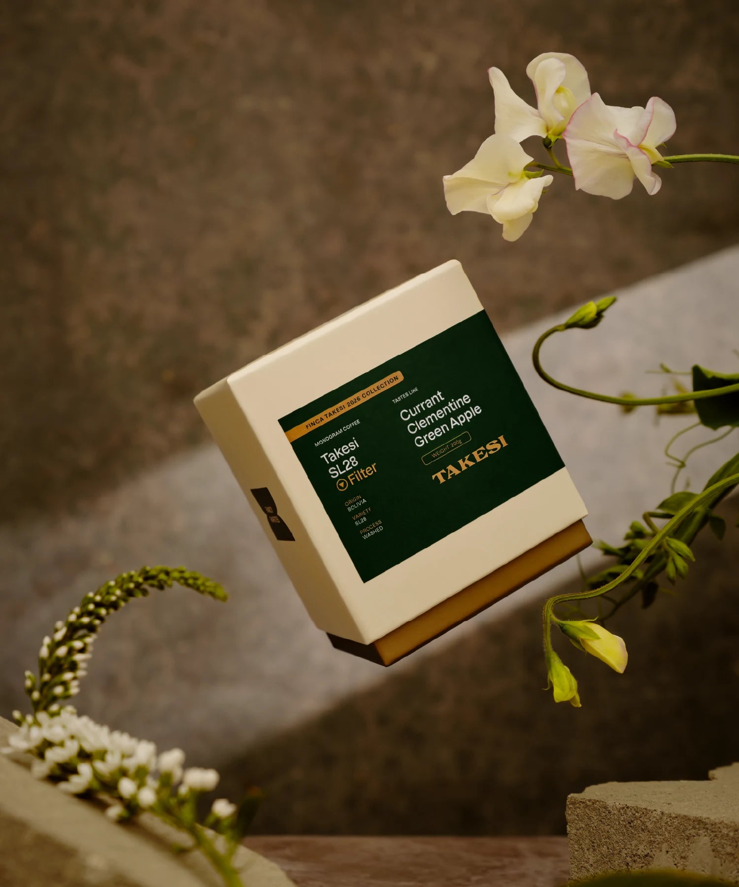 A cream and dark forest green box of Monogram Takesi SL28 filter coffee floating in mid-air, a washed specialty coffee from Bolivia with tasting notes of currant, clementine, and green apple.