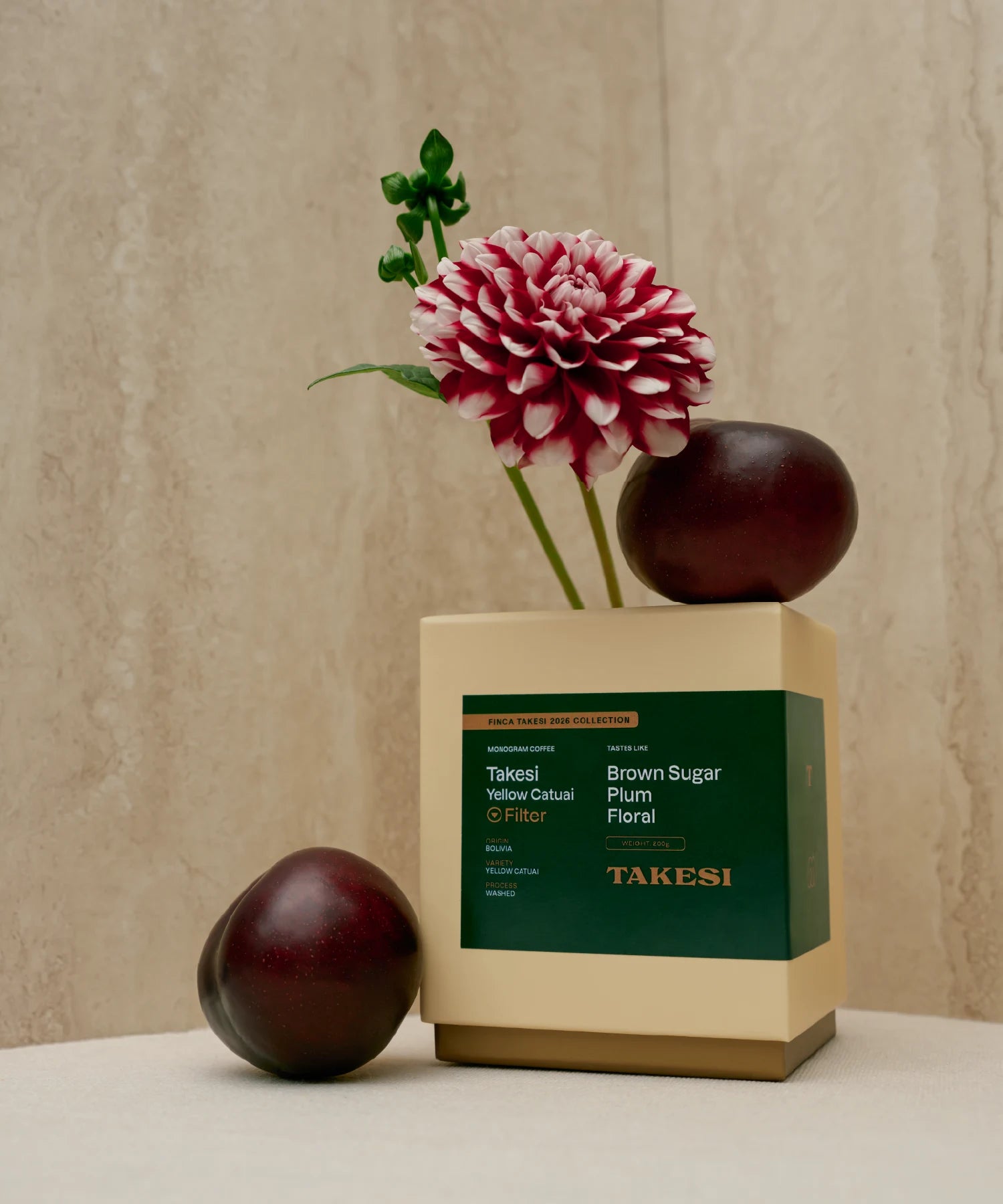 A cream and dark forest green box of Monogram Takesi Yellow Catuai filter coffee in a still life setting, balanced with a deep red plum resting on top next to a striped dahlia flower stem, with a second plum sitting to the left.