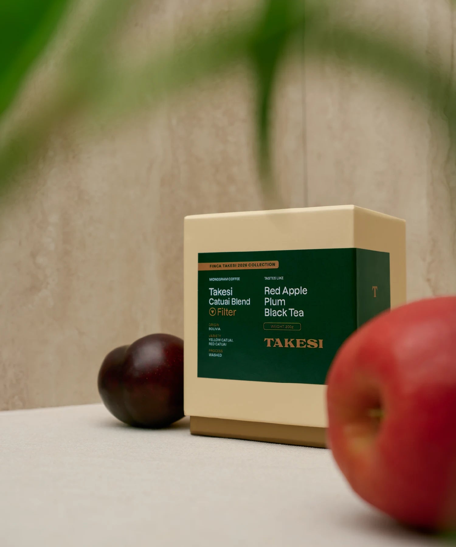 A cream and dark forest green box of Monogram Takesi Catuai Blend filter coffee sitting on a surface in a still life setup, framed by a blurred red apple in the right foreground and a dark red plum resting on the left background.