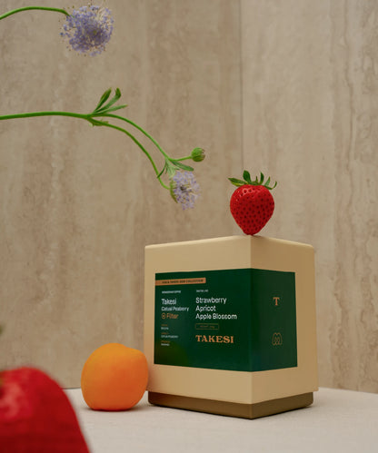 A cream and dark forest green box of Monogram Takesi Catuai Peaberry filter coffee displayed in a still life setting with a fresh strawberry resting on top, an apricot next to it, and a delicate blue flower stem leaning overhead.
