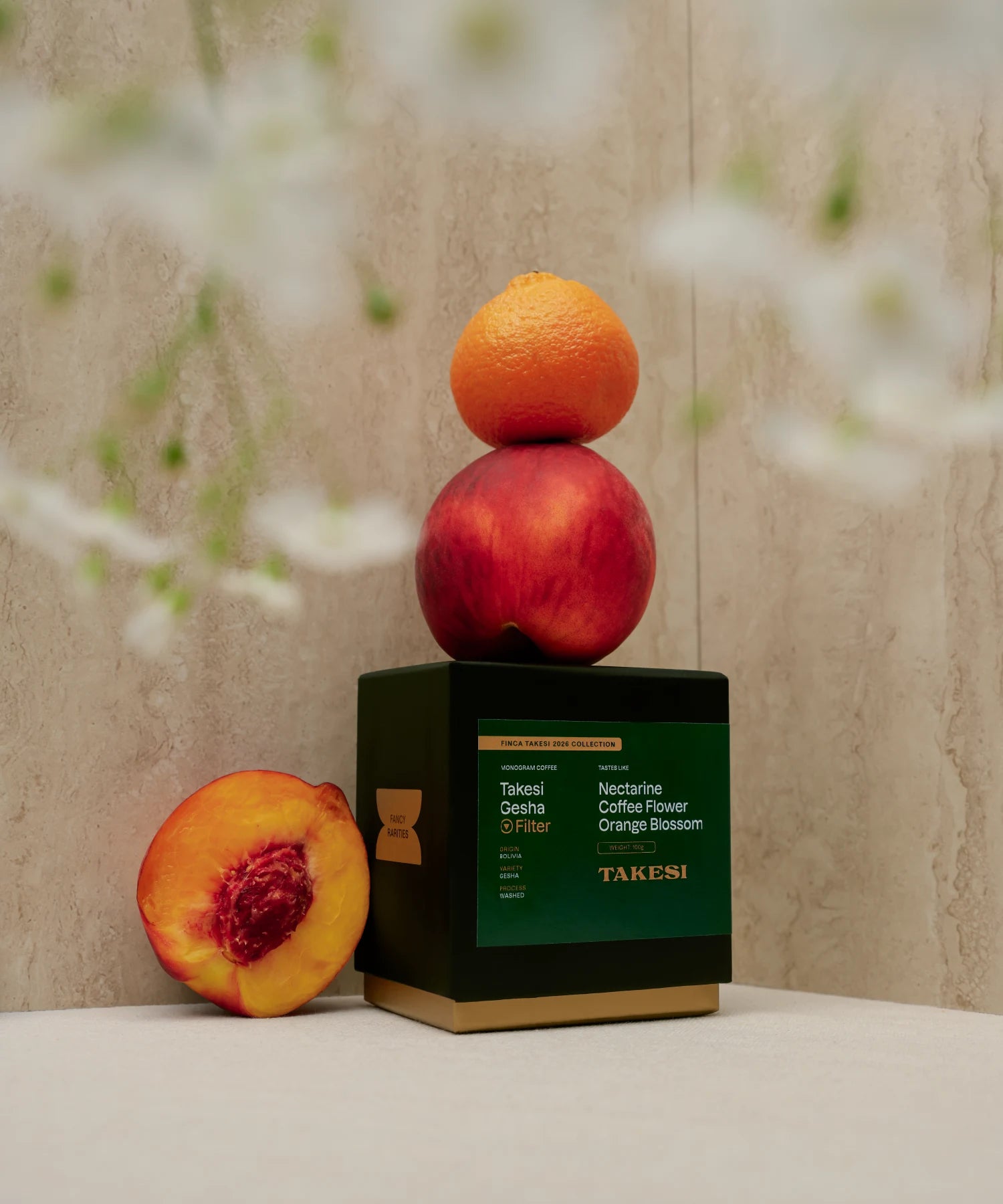 A premium black box with a dark forest green label of Monogram Takesi Gesha filter coffee in a still life setting, stacked vertically with a fresh nectarine and a small orange on top, next to a sliced nectarine showing its pit.