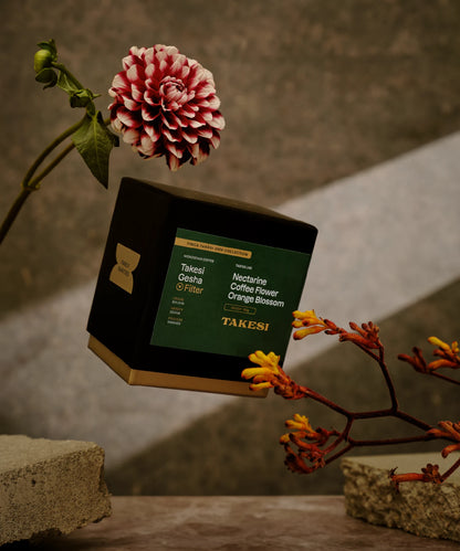 A premium black box with a dark forest green label of Monogram Takesi Gesha filter coffee floating in mid-air, a washed specialty coffee from Bolivia with tasting notes of nectarine, coffee flower, and orange blossom.