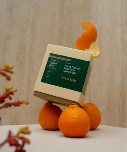 A cream and dark forest green box of Monogram Takesi Java filter coffee dynamically balanced atop three fresh mandarin oranges, with a curled orange peel resting on top against a textured wall backdrop.