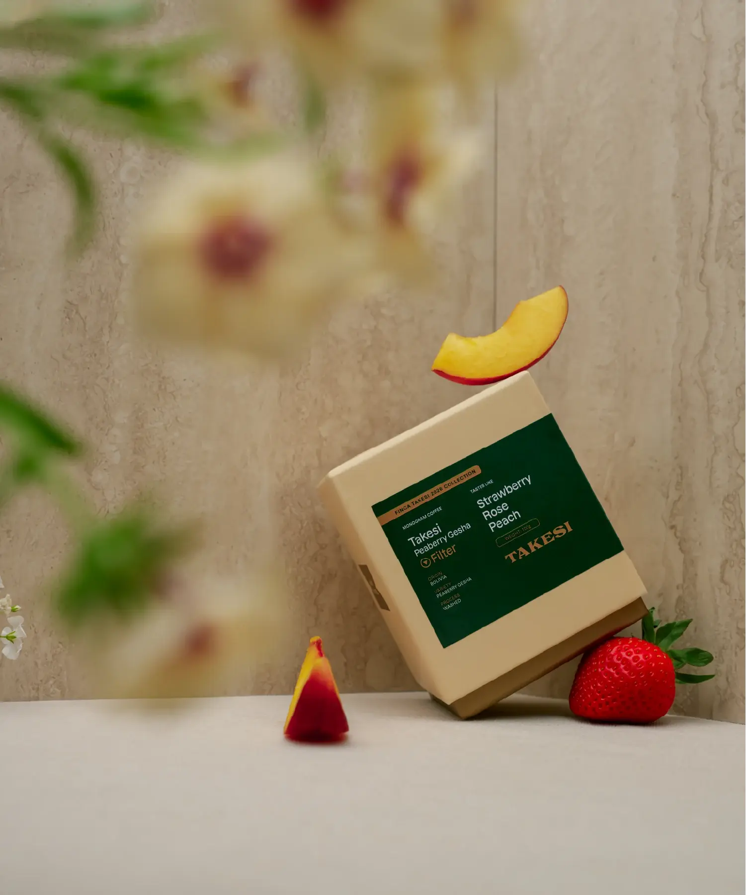 A cream and dark forest green box of Monogram Takesi Peaberry Gesha filter coffee displayed in a still life setting, balanced with a fresh strawberry next to it, a peach slice resting on top, and blurred cream flowers elegantly framing the foreground.