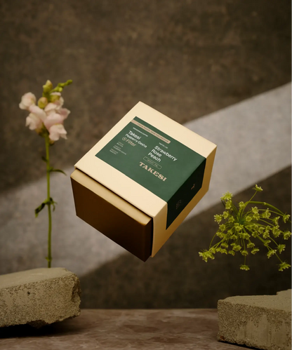 A cream and dark forest green box of Monogram Takesi Peaberry Gesha filter coffee floating in mid-air, a washed specialty coffee from Bolivia with tasting notes of strawberry, rose, and peach.