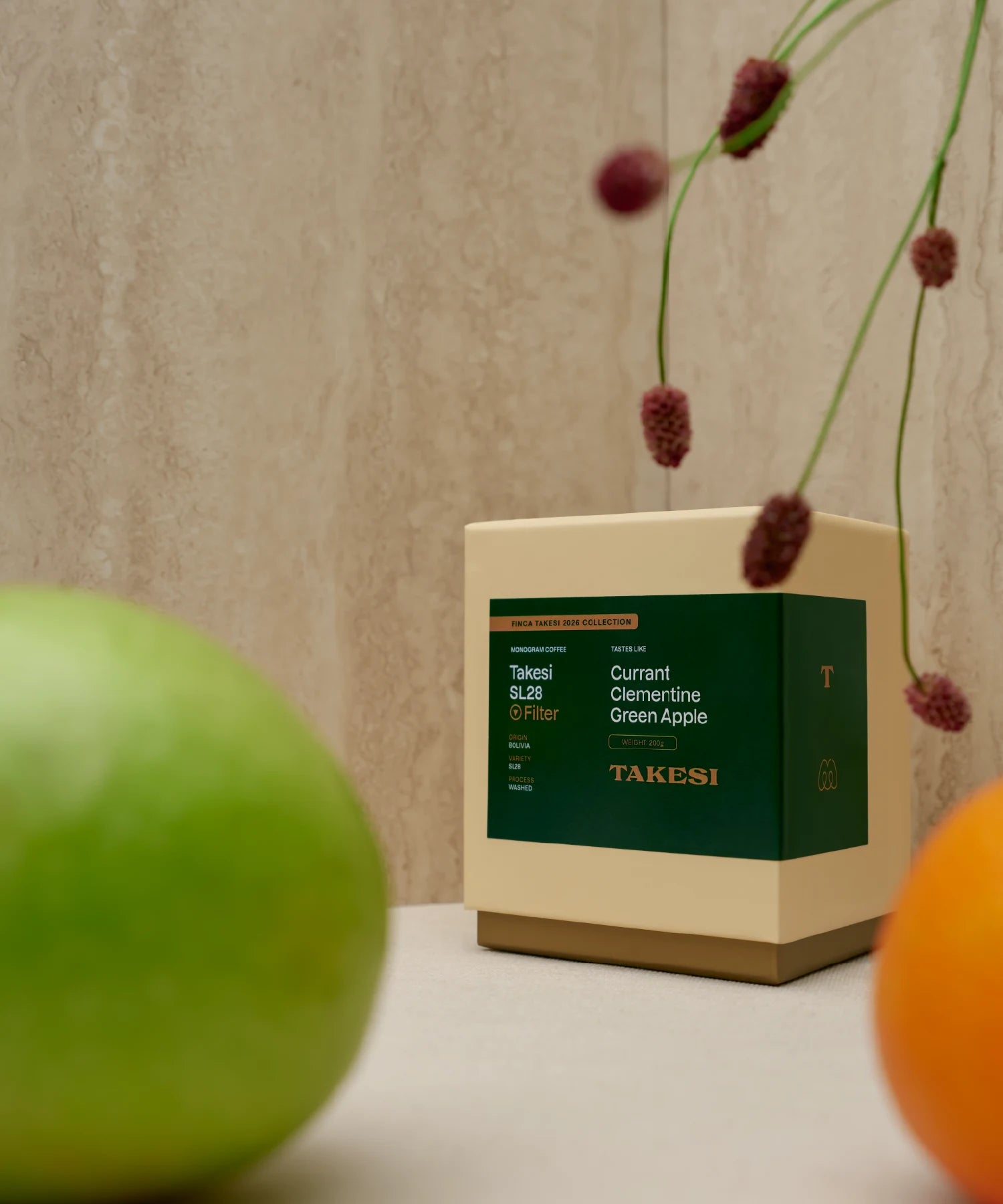 A cream and dark forest green box of Monogram Takesi SL28 filter coffee sitting on a surface in a still life setup, framed by a blurred green apple in the foreground, a mandarin orange on the right, and deep red floral stems overhead