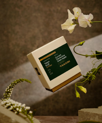 A cream and dark forest green box of Monogram Takesi SL28 filter coffee floating in mid-air, a washed specialty coffee from Bolivia with tasting notes of currant, clementine, and green apple.